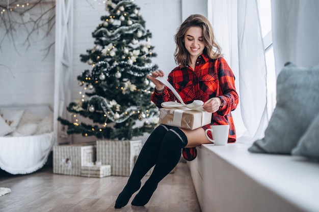 Natale. Bella donna in camicia e calzini seduto sul davanzale della finestra con scatola regalo di Natale, albero di Natale dietro