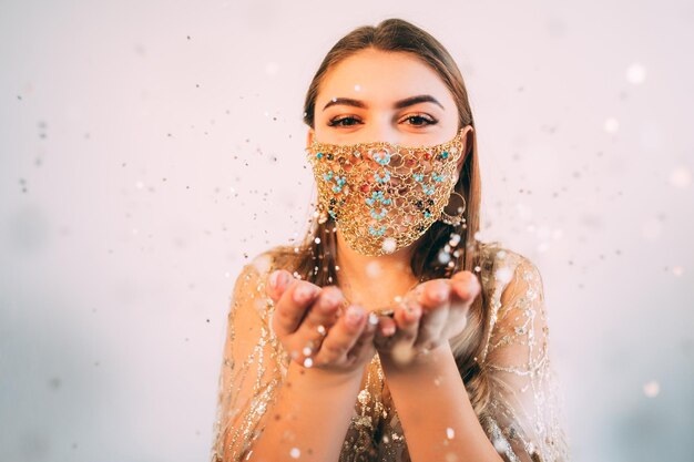 Natale Covid-19. Congratulazioni per le vacanze. Celebrazione delle vacanze invernali in quarantena. Donna felice in maschera facciale a catena d'oro che soffia luce bokeh brilla dalle mani palmi aperti su sfondo rosa spazio vuoto.