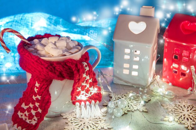 Natale e Capodanno accogliente composizione per le vacanze con tazze bianche con cacao o cioccolato e marshmallow, sciarpa di natale, case di candele in ceramica rossa e bianca sullo sfondo delle luminarie blu del boke