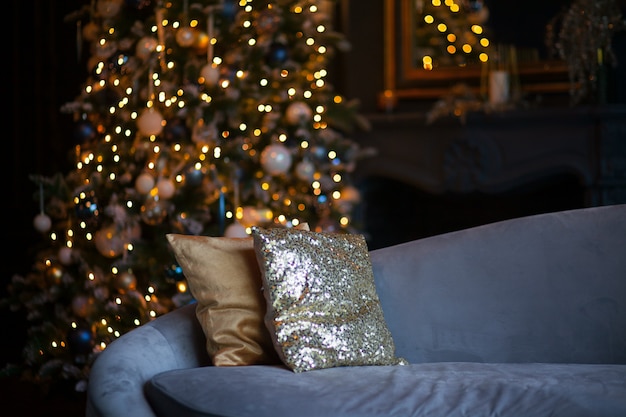 Natale e capodanno hanno decorato la stanza interna scura. Camera arredata per le vacanze con divano azzurro e cuscini dorati. Notte di Natale festivo con luci sull'albero. Notte romantica per due