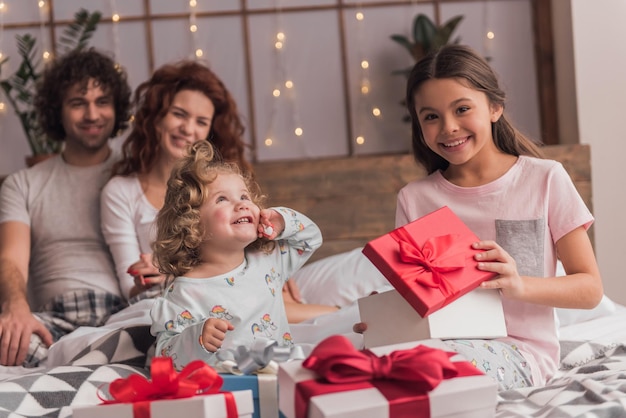 È Natale! Famiglia felice seduta a letto, i genitori sorridono mentre le figlie aprono i regali