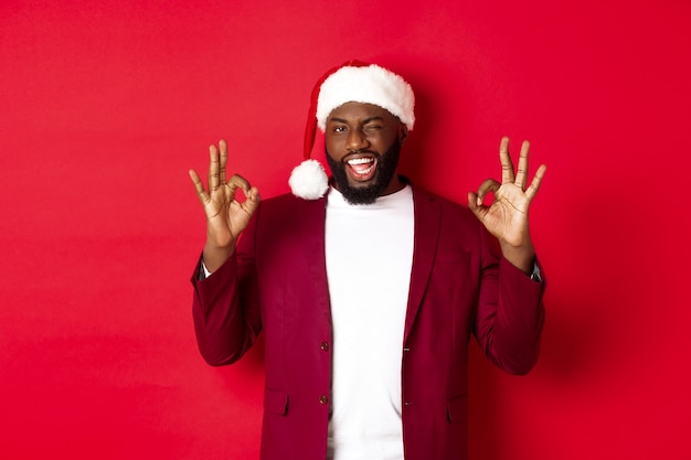 Natale, festa e concetto di vacanze. Bell'uomo afroamericano in cappello della santa, strizzando l'occhio e mostrando segni ok, in piedi su sfondo rosso.