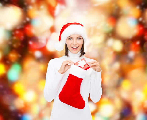 Natale, inverno, felicità, vacanze e concetto di persone - donna sorridente con cappello da Babbo Natale con piccola confezione regalo e calza su sfondo a luci rosse