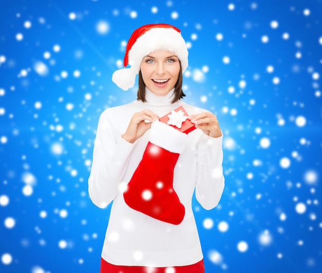 Natale, inverno, felicità, vacanze e concetto di persone - donna sorridente con cappello da Babbo Natale con piccola confezione regalo e calza su sfondo blu innevato