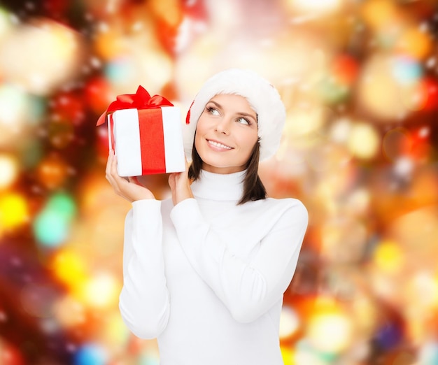 Natale, inverno, felicità, vacanze e concetto di persone - donna sorridente in cappello di Babbo Natale con confezione regalo su sfondo a luci rosse