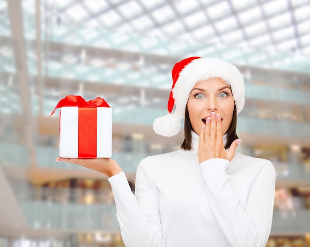 Natale, inverno, felicità, vacanze e concetto di persone - donna sorridente in cappello di Babbo Natale con confezione regalo su sfondo centro commerciale