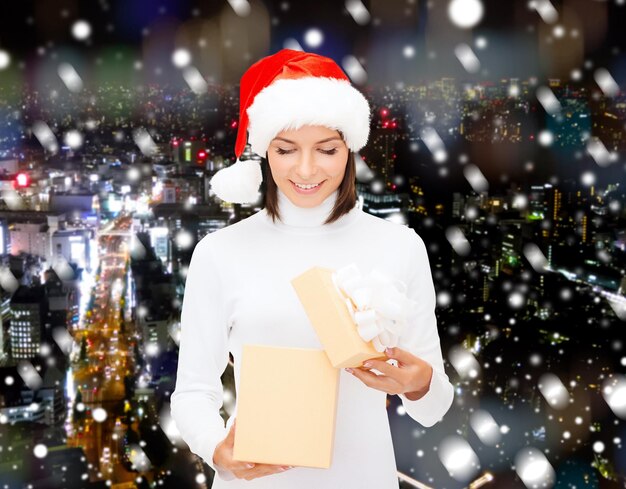 Natale, inverno, felicità, vacanze e concetto di persone - donna sorridente in cappello di Babbo Natale con confezione regalo su sfondo di città di notte innevata