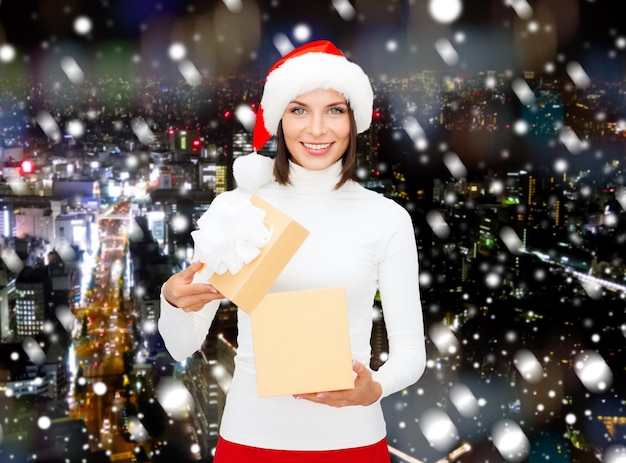 Natale, inverno, felicità, vacanze e concetto di persone - donna sorridente in cappello di Babbo Natale con confezione regalo su sfondo di città di notte innevata