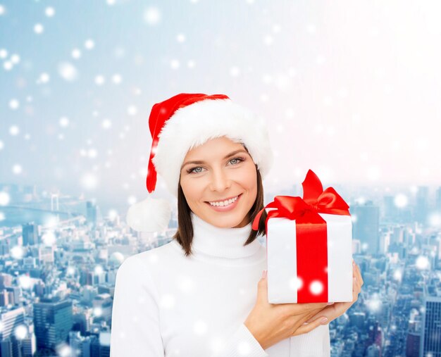 Natale, inverno, felicità, vacanze e concetto di persone - donna sorridente in cappello di Babbo Natale con confezione regalo su sfondo di città innevata