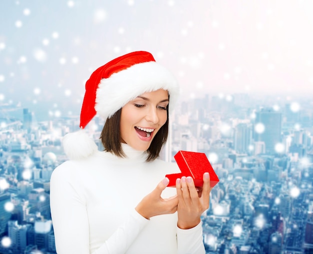 Natale, inverno, felicità, vacanze e concetto di persone - donna sorridente in cappello di Babbo Natale con confezione regalo su sfondo di città innevata