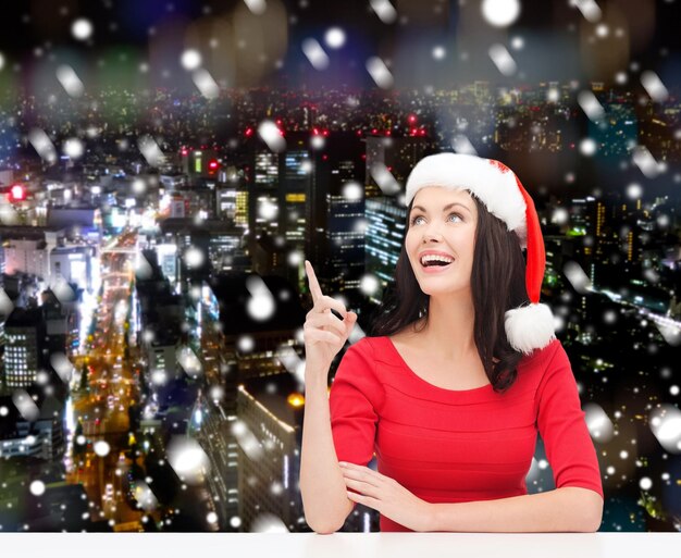 Natale, inverno, vacanze, felicità e concetto di persone - donna sorridente in cappello santa helper che punta il dito su sfondo nevoso della città di notte