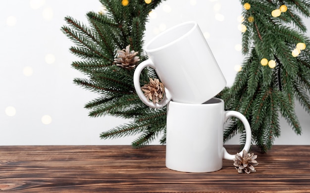 Natale mockup due tazze da tè vuote bianche su un tavolo di legno marrone e rami di una corona di albero di Natale. Modello di tazza per il tuo design, logo con spazio di copia. Capodanno