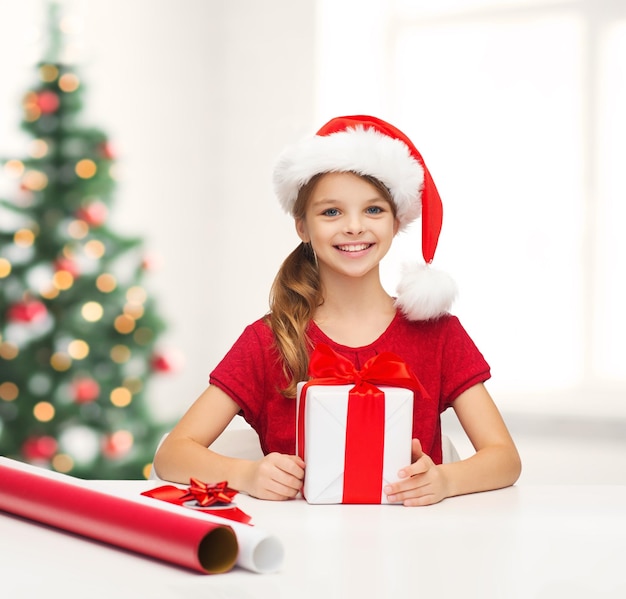 natale, natale, inverno, concetto di felicità - ragazza sorridente con cappello da Babbo Natale con scatola regalo e carta da imballaggio