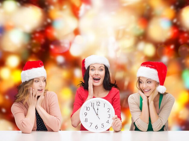 natale, natale, inverno, concetto di felicità - tre donne sorridenti in cappelli di santa helper con orologio che mostra 12