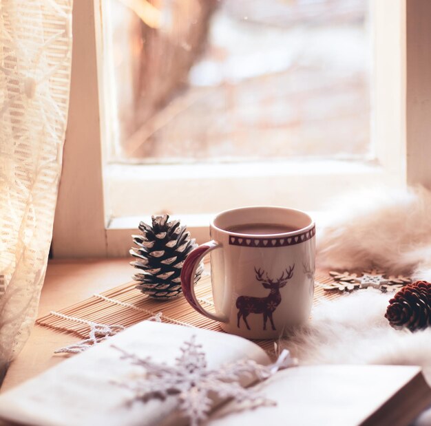 natale natura morta, tazza di cacao, libro, pigne, coperta, fiocchi di neve lavorati a maglia vicino alla finestra luminosa