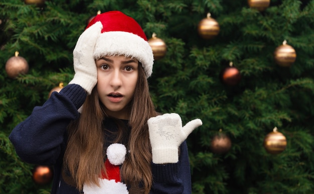 Natale scioccato o sorpreso. Close up Ritratto di donna che indossa un cappello di Babbo Natale con emozione. Sullo sfondo di un albero di Natale.