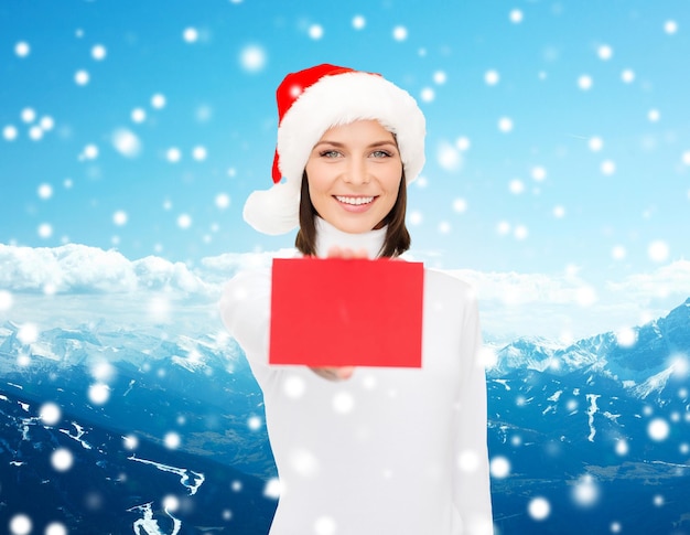 Natale, soggiorni, persone, pubblicità e concetto di vendita - donna felice con cappello da assistente di Babbo Natale con cartellino rosso vuoto su sfondo di montagne innevate