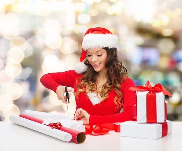 Natale, vacanze, celebrazione, decorazione e concetto di persone - donna sorridente in cappello di Babbo Natale con le forbici che imballano la confezione regalo su sfondo di luci
