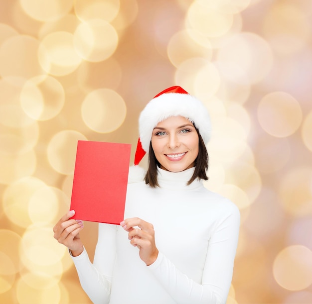 Natale, vacanze, celebrazione, saluto e concetto di persone - donna sorridente in cappello santa helper con biglietto di auguri su sfondo di luci beige