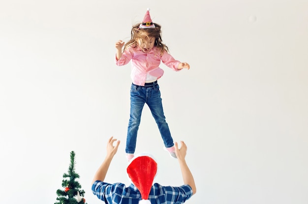 Natale, vacanze e concetto di paternità - Papà divertente che indossa il cappello di Babbo Natale lancia sua figlia su sfondo bianco con spazio di copia.