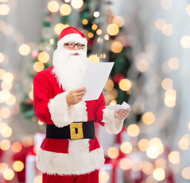 Natale, vacanze e concetto di persone - uomo in costume di Babbo Natale che legge la lettera su sfondo di luci ad albero