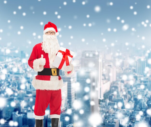 Natale, vacanze e concetto di persone - uomo in costume di babbo natale con confezione regalo che mostra pollice in alto gesto su sfondo città innevata