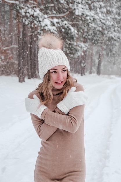 Natale, vacanze e concetto di stagione. Giovane donna felice che soffia neve nella natura della foresta invernale. Abbigliamento caldo guanti e cappello lavorati a maglia. Sfondo di paesaggio forestale invernale