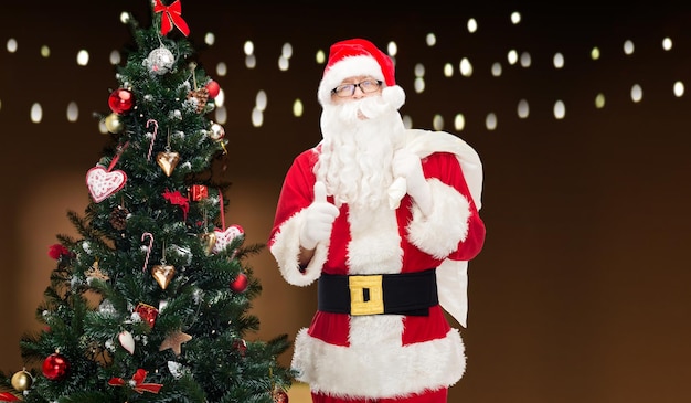 Natale, vacanze, gesto e concetto di persone - uomo in costume di Babbo Natale con borsa e albero di Natale che mostra i pollici su sfondo di luci di ghirlande