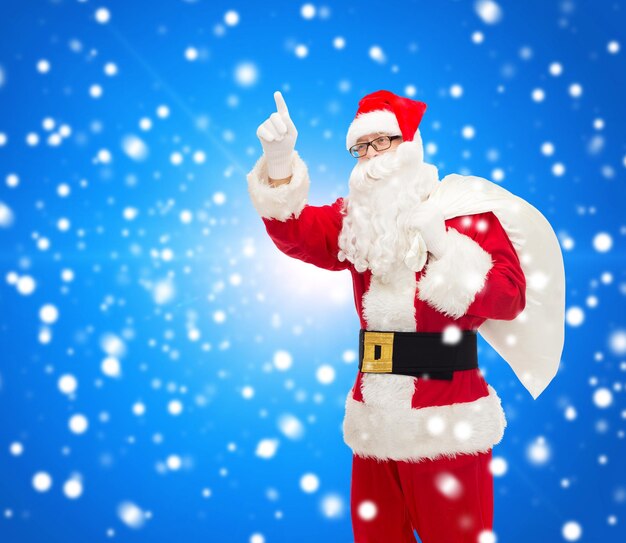 Natale, vacanze, gesto e concetto di persone - uomo in costume di babbo natale con la borsa che punta il dito su sfondo blu innevato