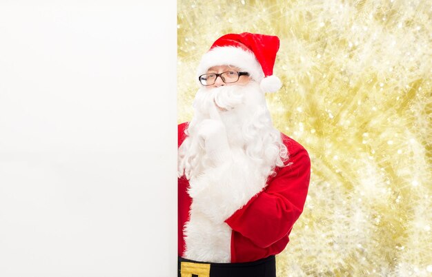 Natale, vacanze, pubblicità e concetto di persone - uomo in costume di babbo natale con cartellone bianco bianco che fa un gesto silenzioso su sfondo a luci gialle