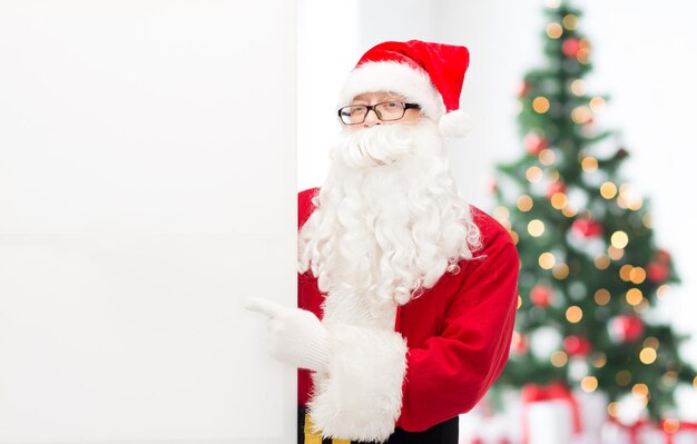 Natale, vacanze, pubblicità, gesto e concetto di persone - uomo in costume di babbo natale che punta il dito su un cartellone bianco bianco sopra il soggiorno con albero