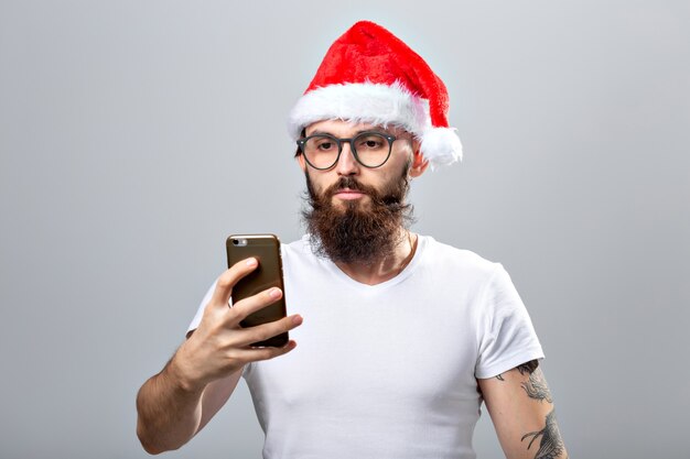 Natale, vacanze, tecnologia e concetto di persone - bell'uomo barbuto con cappello santa che scatta foto selfie con smartphone su sfondo grigio.