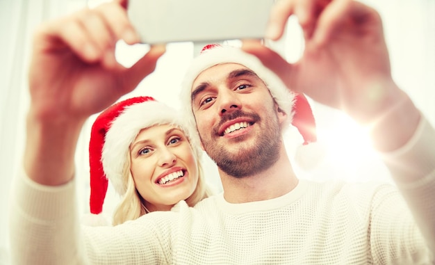 Natale, vacanze, tecnologia e concetto di persone - coppia felice con cappelli di Babbo Natale che si fa un selfie con lo smartphone a casa