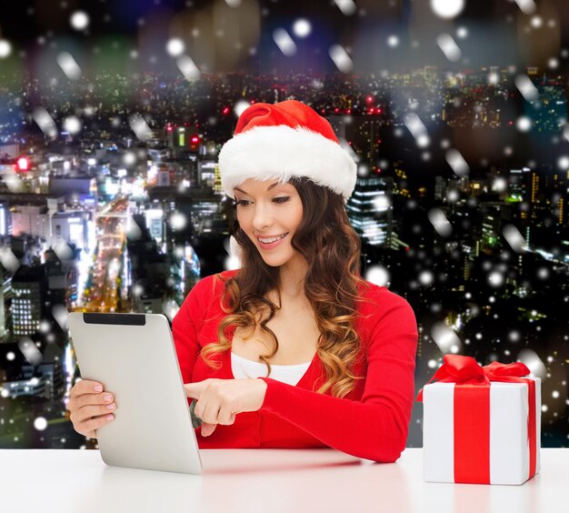 Natale, vacanze, tecnologia e concetto di persone - donna sorridente con cappello da assistente di Babbo Natale con confezione regalo e computer tablet pc su sfondo nevoso della città di notte