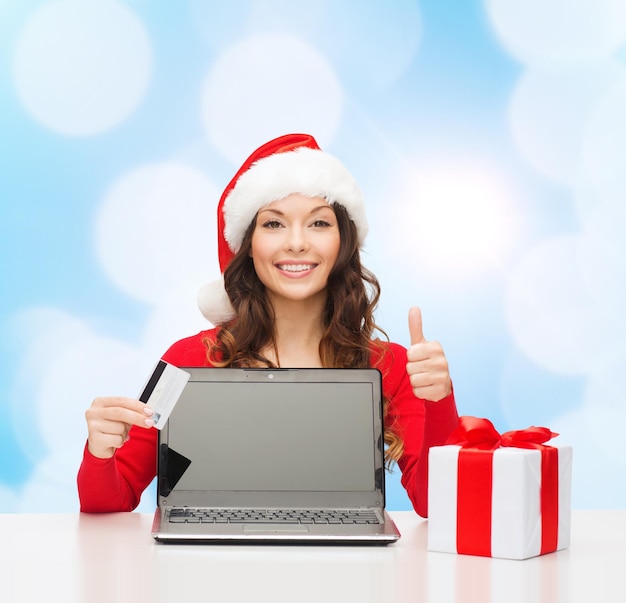 Natale, vacanze, tecnologia e concetto di shopping - donna sorridente con cappello da Babbo Natale con carta di credito, confezione regalo e computer portatile che mostra il pollice in alto gesto su sfondo di luci blu