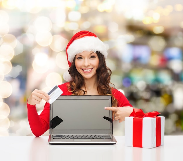 Natale, vacanze, tecnologia e concetto di shopping - donna sorridente con cappello da Babbo Natale con confezione regalo, carta di credito e computer portatile che punta il dito su sfondo di luci