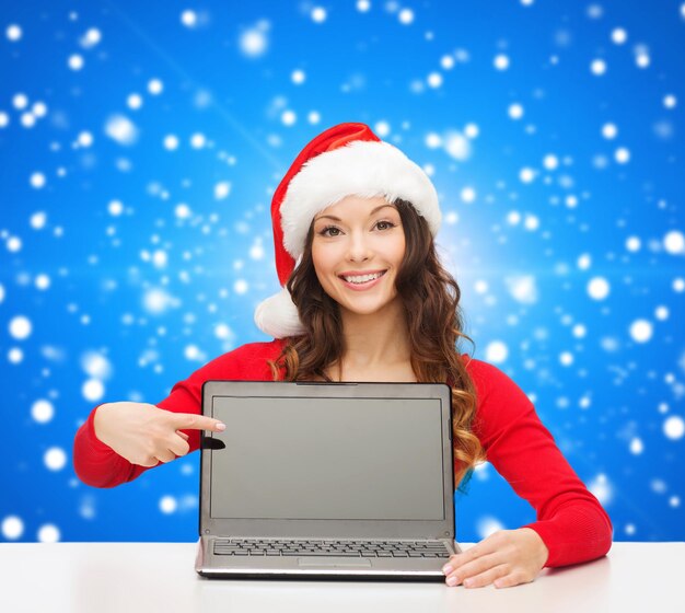 Natale, vacanze, tecnologia, pubblicità e concetto di persone - donna sorridente in santa helper hat pointig dito sullo schermo del computer portatile vuoto su sfondo blu nevica