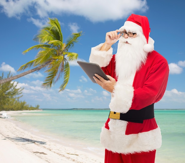 natale, vacanze, viaggi, tecnologia e concetto di persone - uomo in costume di babbo natale con computer tablet pc su sfondo spiaggia tropicale