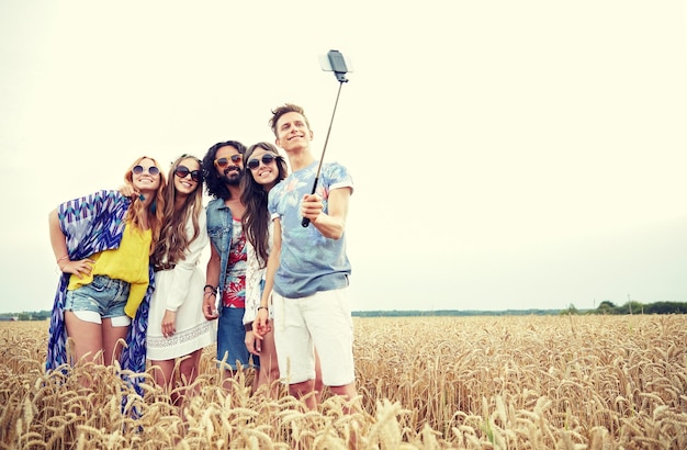 natura, estate, cultura giovanile, tecnologia e concetto di persone - giovani amici hippie sorridenti in occhiali da sole che scattano foto con lo smartphone sul bastone selfie sul campo di cereali