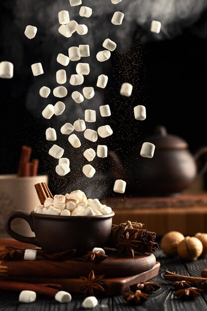 Natura morta con cioccolata calda e marshmallow. Marshmallow e cannella in polvere cadono dall'alto. Il vapore caldo sale dalla tazza