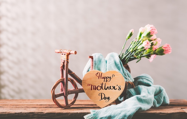 Natura morta con fiori di garofano dolce in bicicletta di legno sul tavolo di legno, concetto di giorno di madri con un messaggio per la festa della mamma felice su un cuore di legno