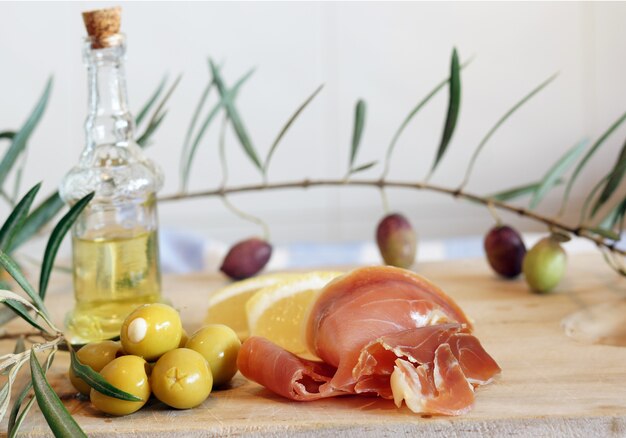 Natura morta con prosciutto spagnolo a pata negra, olive e olio d'oliva in bottiglia su un tagliere di legno.