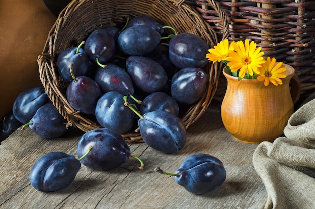 Natura morta con prugne mature in un cesto e fiori gialli in stile rustico. ¡perdere. Colpo verticale.