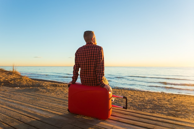 Natura, viaggi e concetto di persone. vista posteriore dell'uomo seduto su una valigia rossa vicino al mare e guardando il tramonto