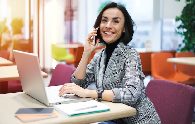 Nel coworking. Foto vista laterale di una donna allegra in abiti casual eleganti, che parla al telefono mentre lavora al suo laptop.