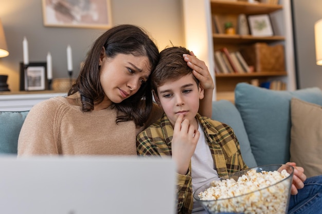 Nel loro soggiorno una madre e suo figlio condividono una serata di film accogliente con i popcorn in mano guardano un film triste sul portatile accovacciati sul divano trovando conforto nella compagnia dell'altro