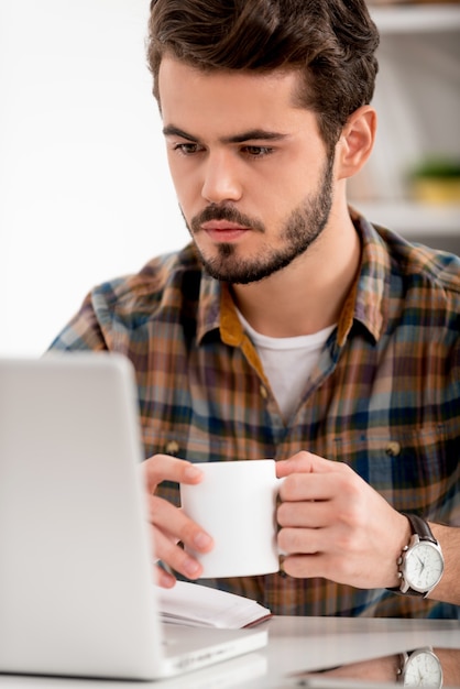 Nel profondo del suo lavoro. Giovane premuroso che guarda il laptop e tiene in mano una tazza di caffè mentre è seduto al suo posto di lavoro