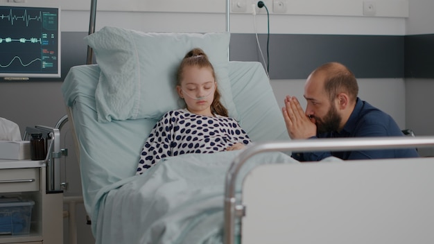 Nel reparto ospedaliero un piccolo paziente che dorme mentre il padre preoccupato prega per la guarigione dalla malattia