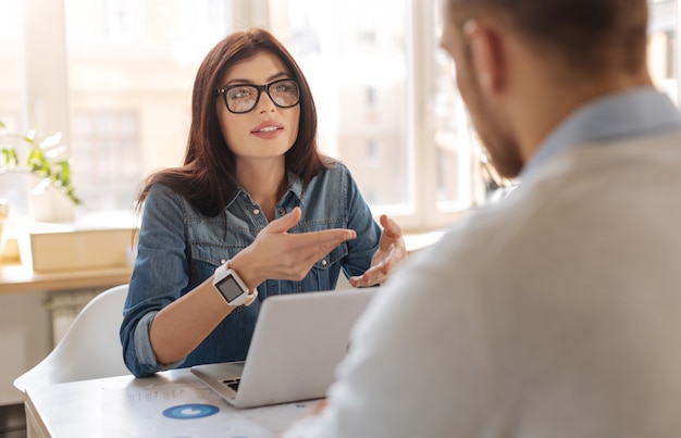 Nell'ufficio. Attraente donna intelligente e piacevole seduta al tavolo e guardando il suo collega mentre gli spiega qualcosa