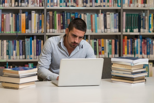 Nella biblioteca Studente maschio bello con il computer portatile ed i libri che lavorano in una profondità del campo poco profonda della biblioteca dell'università della High School